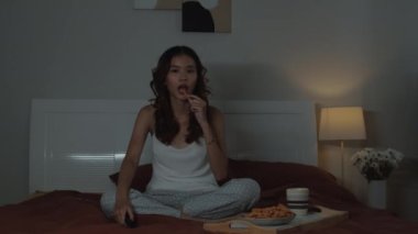 Genç bir kızın gece yarısı tek başına korku filmi izlerken yatakta oturup abur cubur yediği orta boy bir fotoğraf.