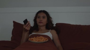 TV programları seyrederken, geceleri cips ya da atıştırmalık yerken, Asyalı etnik kökenli bir kızın yatakta uzanıp yalnız vakit geçirmenin keyfini çıkardığı orta boy bir fotoğraf.