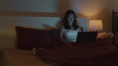 Düşünceli kadın işkoliğin gece yatakta otururken kablosuz bilgisayarla ilgili haber yaparken orta uzunlukta bir görüntüsü.