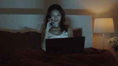 Genç serbest çalışan bir bayanın iş arkadaşını arayıp gece geç saatlere kadar laptopun önünde yatarken yeni projenin detaylarını sorduğu orta boy bir fotoğraf.