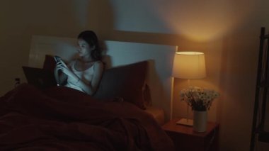Genç Asyalı bir kadının dizüstü bilgisayarla çalışırken cep telefonuna baktığı orta boy bir fotoğraf. Geceleri yatakta oturuyor, uzayı kopyalıyor.