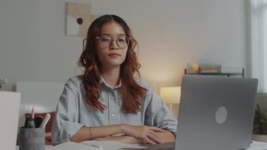 Asyalı kadın portresi kablosuz bilgisayarın önünde oturuyor ve evden kameraya bakıyor.