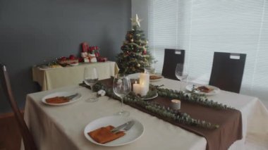 Şarap kadehleri, tabaklar, çatal bıçaklar ve mumlarla dolu el kamerasıyla Noel ağacı ve arka planda hediyeler.