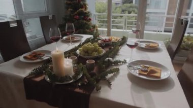 Noel kutlaması için dekore edilmiş rahat oturma odasının orta uzunluktaki görüntüsü farklı mezeler, şarap ve yanan mumlarla dolu festival masası arka planda yeni yıl ağacı.