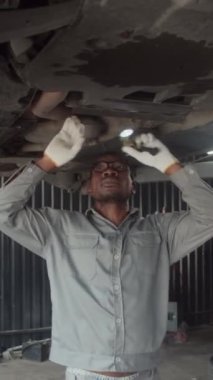 Arabanın altını tamir ederken gri üniformalı Afrikalı Amerikalı tamircinin el feneri kullanırken dikey görüntüsü.