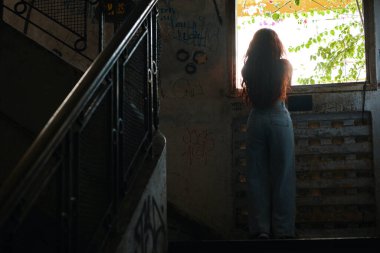 Uzun saçlı genç bir kadın terk edilmiş ve çürüyen bir binanın penceresine bakıyor.