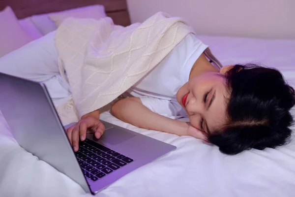 Sleepy girl lying on bed under throw blanket and watching movie on laptop or video calling friend