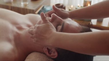 Spa salonundaki güzellik prosedürü sırasında rahatlamış bir adamın yaş karşıtı yüz masajı yaptırışının yan görüntüsü