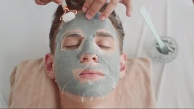 Melez bir gencin gözleri kapalı bir şekilde kanepede uzanması ve güzellik prosedürü ile kaplıca stüdyosunda maske takması.