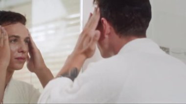 Genç ve çekici bir adamın banyoda aynanın önünde yüzüne nemlendirici krem sürerken.