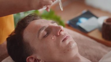 Kadın uzmanın, kuaförde güzellik prosedürü yapan etnik kökenli genç bir adamın derisine yüz serumu eklemesine yakın çekim.