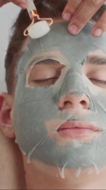 Yukarıdan bakıldığında, genç bir adam kanepede yatarken, spa stüdyosunda maske ve yeşim taşıyla yüz bakımı yaptırıyor.