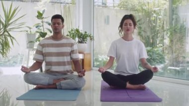 Çift ırklı çiftin oturma odasında oturma odasında panoramik pencerelerle lotus pozu verirken yoga çalışmalarını çekiyoruz.