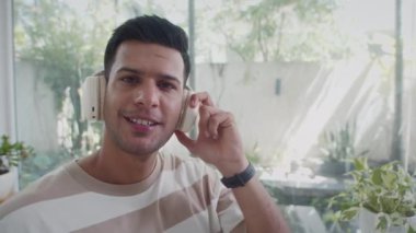 Kulaklık takan, müzik dinleyen ve evde kamera izleyen genç Hintli adamın portresi.