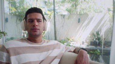 Ev elbiseli genç Hintli bir adamın rahat oturma odasında kulaklıkla müzik dinlemesi.