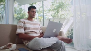 Ev elbiseli bir işadamının rahat oturma odasındaki panoramik pencereli kanepede otururken kablosuz bilgisayarla çalıştığı orta boy bir fotoğraf.