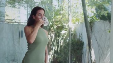 Hafta sonunu evde tek başına geçirirken yeşil elbiseli, bir fincan bitki çayı içen çekici bir kadının orta boy fotoğrafı.