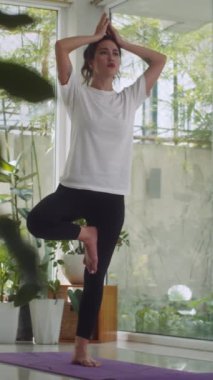Beyaz kadın dikey olarak spor kıyafetleri içinde ağaç duruşu yapıyor ve evde yoga yapıyor.