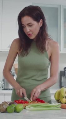 Yeşil elbiseli, salata için sebze doğrayan ve mutfakta yemek tarifine bakan beyaz bir kadının dikey görüntüsü.