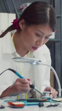 Servis merkezinde bozuk telefonu tamir ederken lehim demiri kullanan beyaz önlüklü kadın teknisyen dikey.