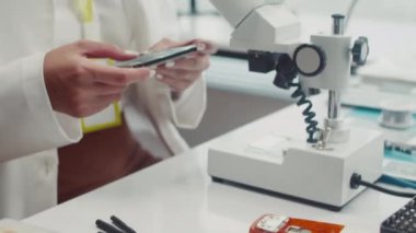 Beyaz önlüklü kadın el kamerasıyla kırılmış cihazın bileşenlerini alıyor ve servis merkezinde mikroskop altında inceliyor.