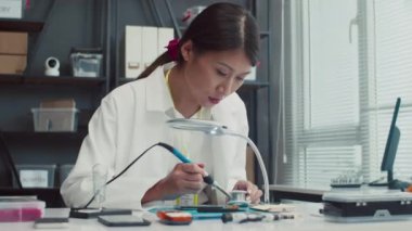 Beyaz işçi kıyafetli bir kadının servis merkezinde bozuk akıllı telefonu tamir ederken lehim demiriyle çalışırken orta boy fotoğrafı.