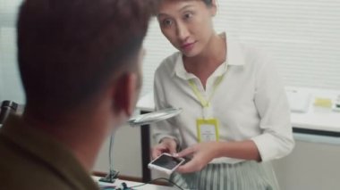Kadın bir uzmanla konuşan bir adamın omzunun üstünden servis merkezinde cep telefonunu inceliyor.