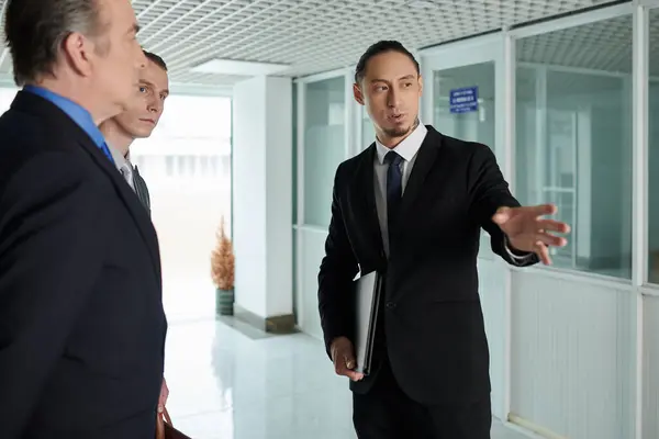 Businessman in black suit meeting business partner in office hall and explaining how to get to boardroom
