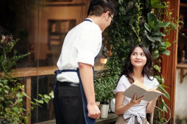 Barista elinde kitapla dışarıda masada oturan genç bayana kahve getiriyor.