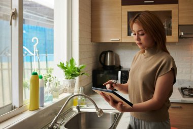 Mutfakta sızdıran musluğu değiştirirken tablet bilgisayardan özel ders izleyen ciddi bir genç kadın.