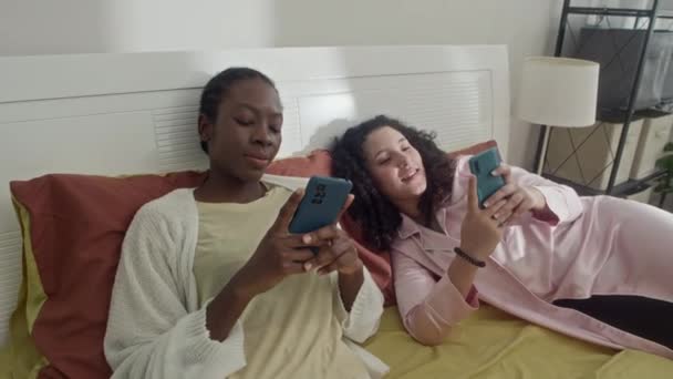 身着睡衣 头脑发热的女性朋友躺在床上 在智能手机上查看社交媒体的中景照片 — 图库视频影像