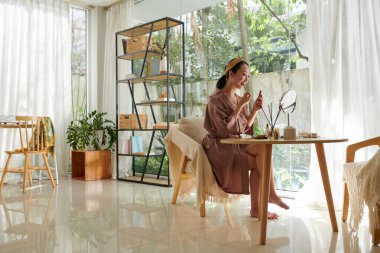 İpek bornozlu genç bir kadın randevuya hazırlanırken makyaj yapıyor.