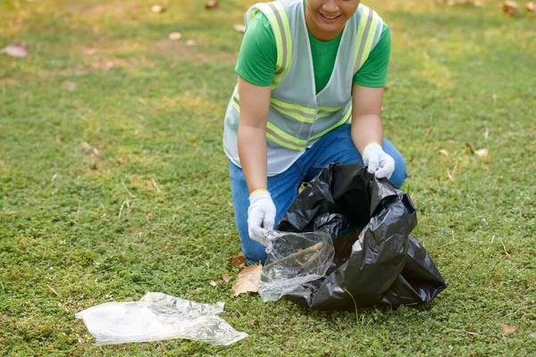 Smiling volunteer in textile gloves picking garbage in local park,