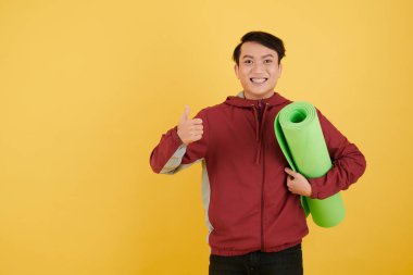 Elinde yoga minderi tutan ve baş parmağını gösteren heyecanlı genç adamın portresi.