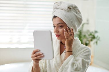 Banyoda kameranın önünde dikilip yüzüne bakarken elinde aynaya bakan bornozlu genç bir kadın.