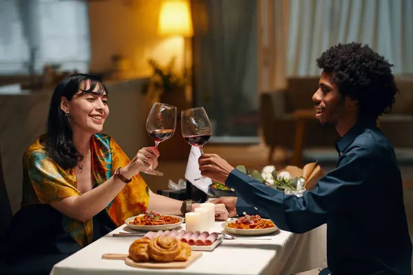 Happy young sweethearts toasting for love and clinking with glasses of red wine while sitting by served festive table and celebrating Valentine Day