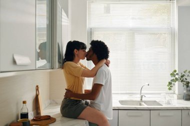 Giyim kuşamında genç bir kadın mutfak tezgahında oturup kocasını öpüyor ve ellerini beline koyuyor.