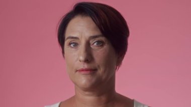 Orta yaşlı, neşeli, mavi gözlü, siyah saçlı, pembe arka planda kameraya gülümseyen bir kadının portresi.