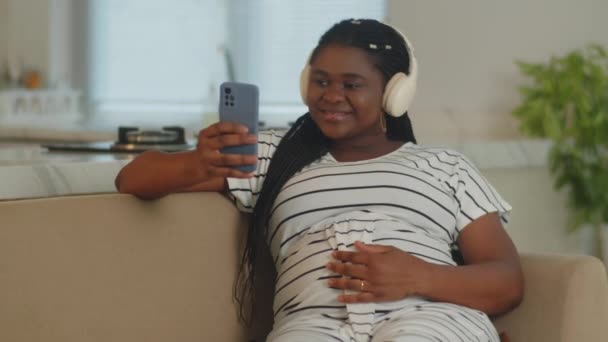 身着条纹衣服 头戴耳机 在家里听音乐时使用智能手机的非洲裔美国孕妇的中景照片 — 图库视频影像