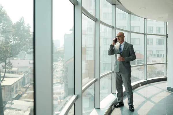 Confident mature chief executive officer with smartphone by ear and cup of coffee in hand standing by large window in office center