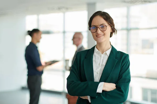 Young successful female chief executive officer in eyeglasses and formalwear looking at camera with smile against two colleagues