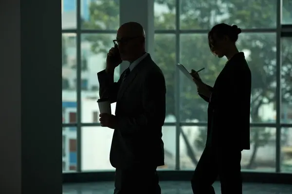 Side view of outlines of two business people standing against large window in office center, speaking on mobile phone and making notes