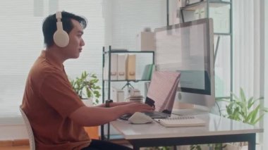 Kulaklıklı neşeli Asyalı kodcunun yan portresi bilgisayarda yazılım uygulaması geliştiriyor ve kameraya bakıyor.