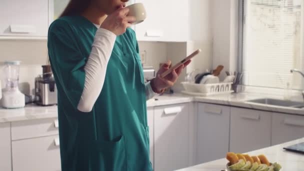 医学の制服飲茶で若い大人の看護師のショットと家庭のキッチンでスマートフォンを使用 — ストック動画