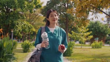 Elinde su ve elma tutan üniformalı neşeli genç bir sağlık görevlisinin yaz parkındaki kliniğe giderken orta boy fotoğrafı.