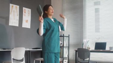 Klinikte sınavı geçtikten sonra hemşire odasında dans eden, üniformalı, neşeli Asyalı bir doktor.