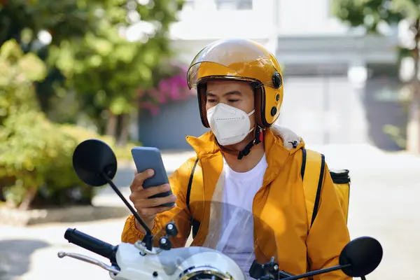 Serious delivery man sitting on motorbike when reading next order notification on smartphone