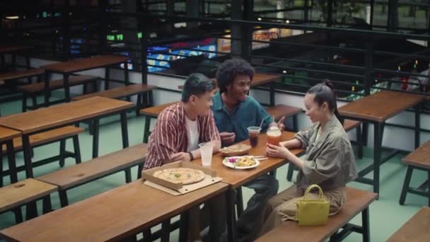 食堂の大きなテーブルに座ってピザやスナックを食べる多文化の友人のワイドショット — ストック動画