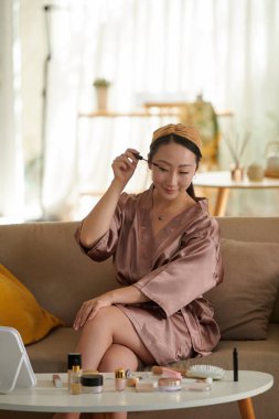 Saten bornozlu gülümseyen kadın sabah hazırlanırken maskara sürüyor.