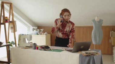 Orta boy Asyalı terzinin cep telefonuyla müşteriyi arayıp atölyedeki elbise detaylarını sorması.